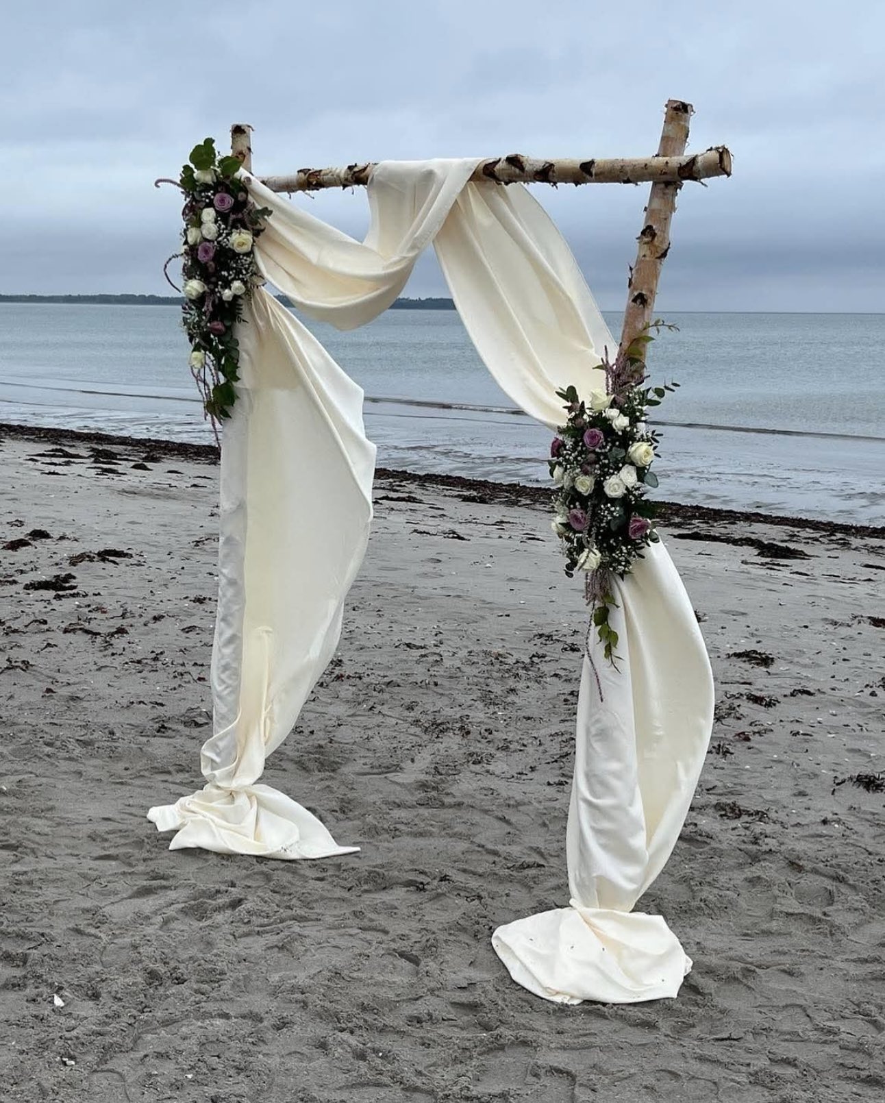 Romantisk blomsterpynt på bryllupsbue ved stranden – med hvide og lilla blomster arrangeret af Fru P